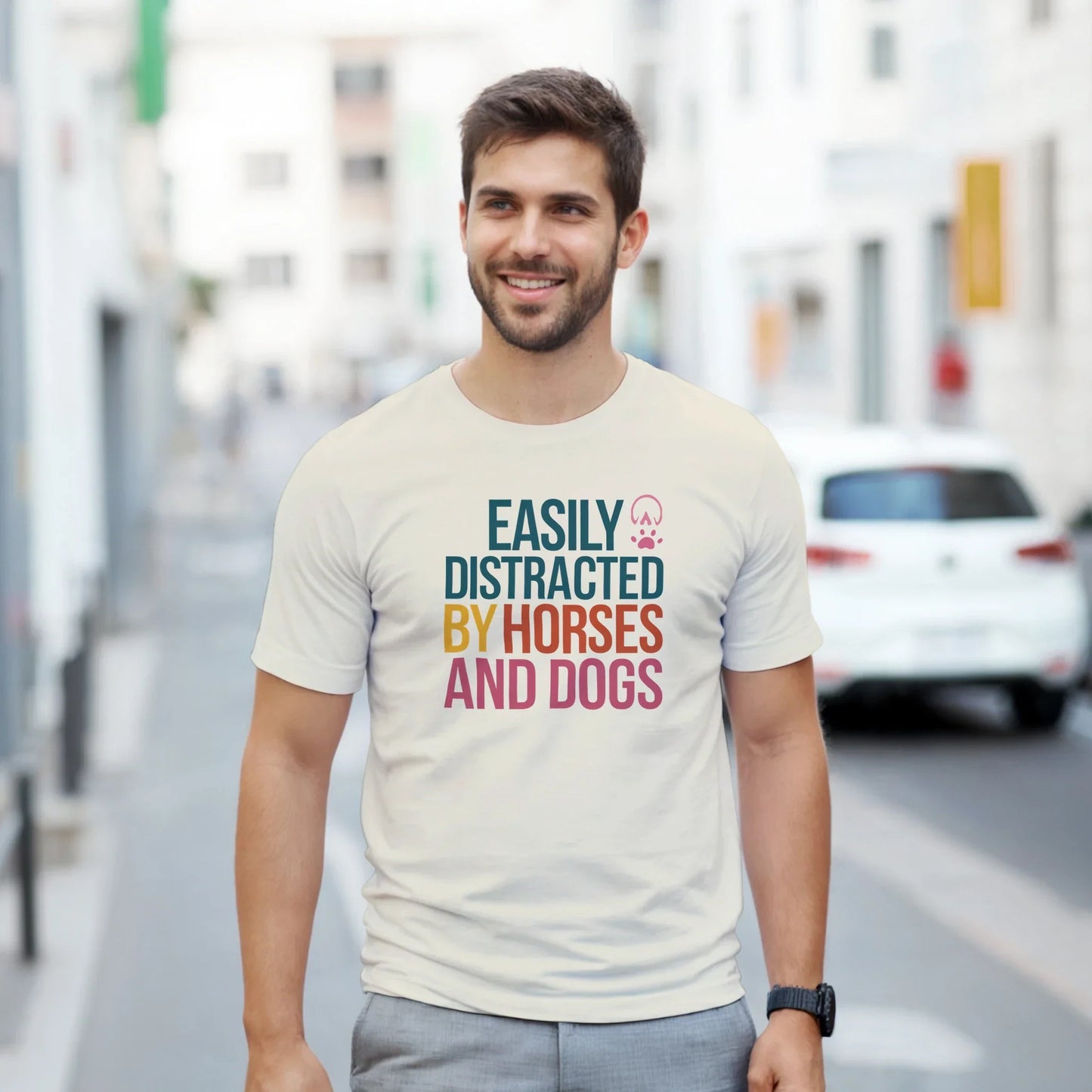 Man wearing Easily Distracted By Horses and Dogs T-Shirt with colorful text on a casual street background