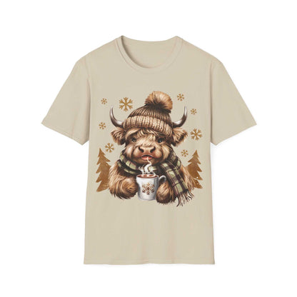 Beige t-shirt featuring a cozy Highland Cow wearing a knit hat and plaid scarf holding a hot chocolate mug with snowflakes.