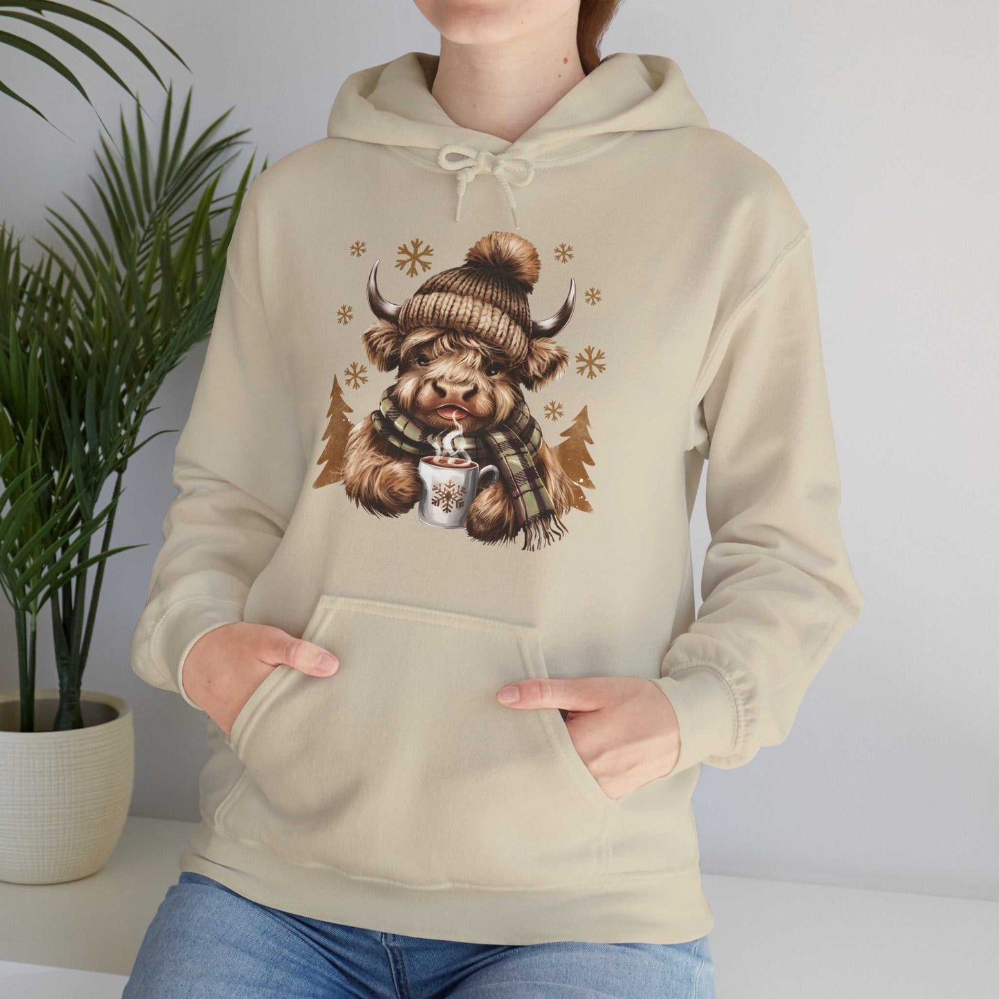Beige Highland Cow hoodie with cozy winter design featuring a cow in a knit hat and scarf holding a hot chocolate mug with snowflakes