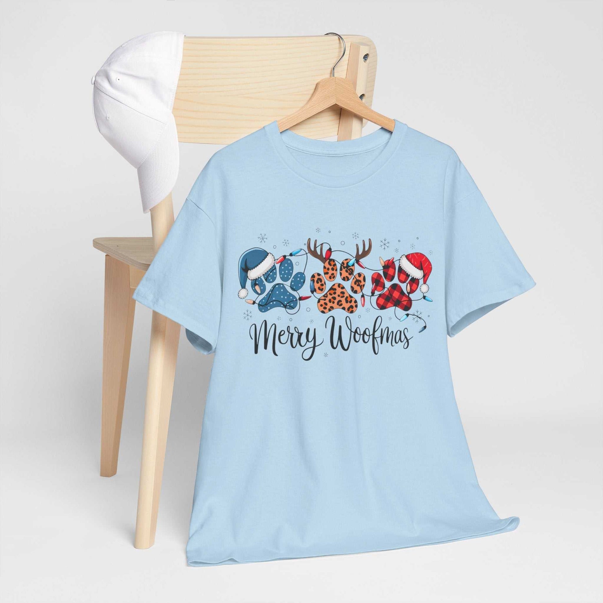 Light blue Merry Woofmas Christmas tee with festive paw prints in buffalo plaid, leopard spots, Santa hats, and antlers displayed on a chair with a white cap.