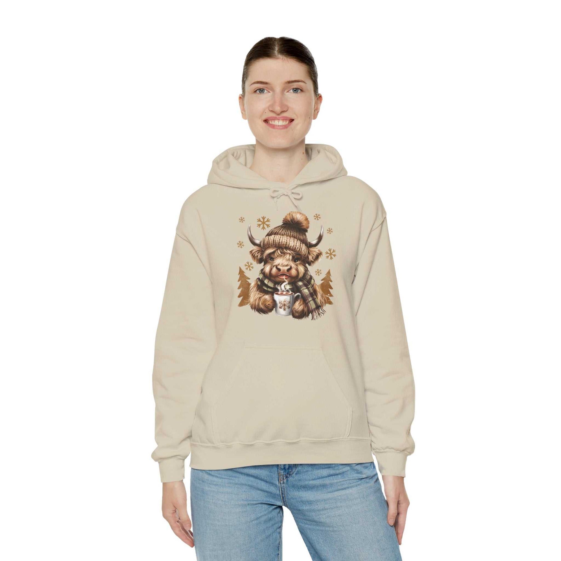 Woman wearing beige Highland Cow Hoodie with winter-themed cow holding hot chocolate in knit hat and plaid scarf design