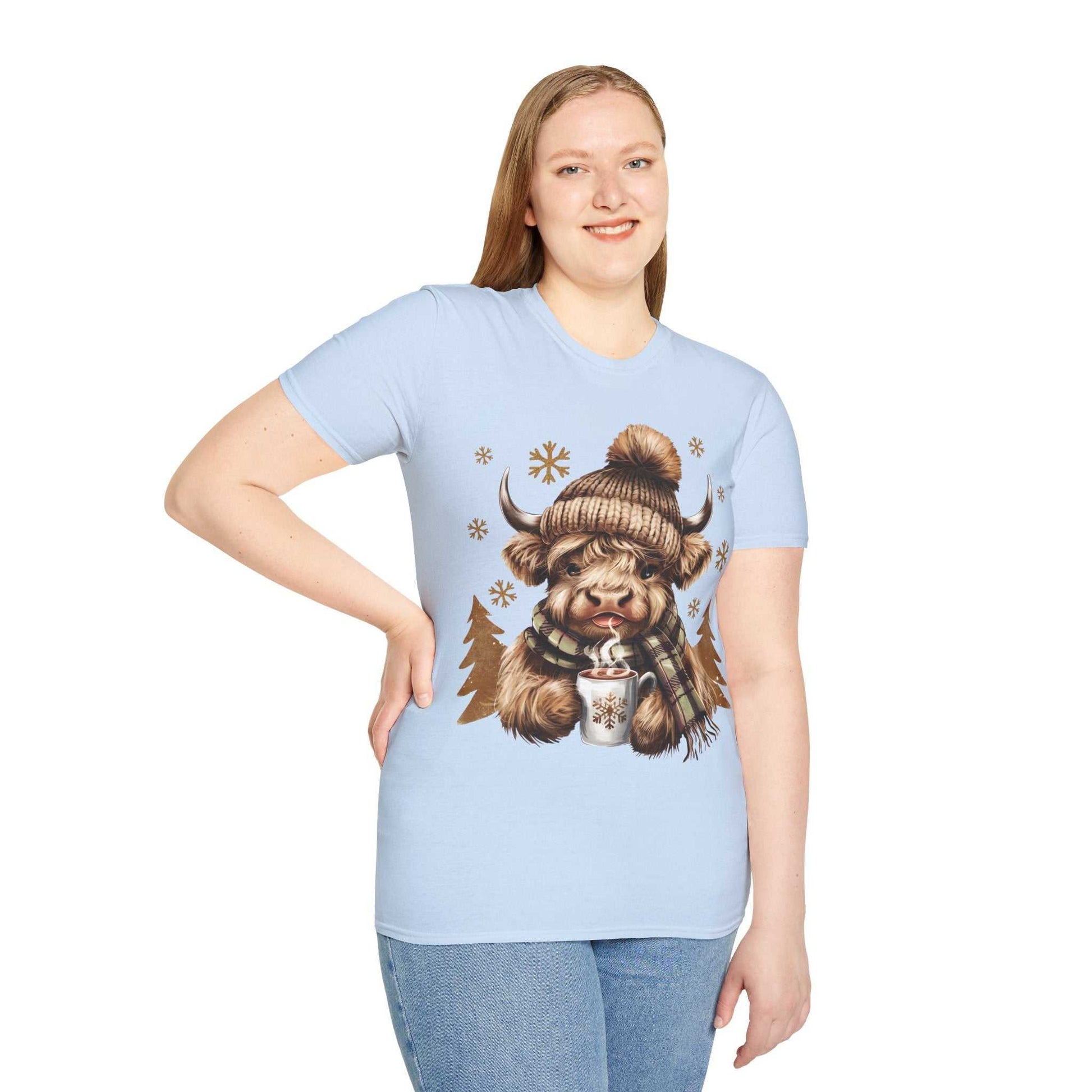 Light blue t-shirt featuring a cozy Highland Cow with a knit hat, plaid scarf, and hot chocolate design, worn by a smiling woman