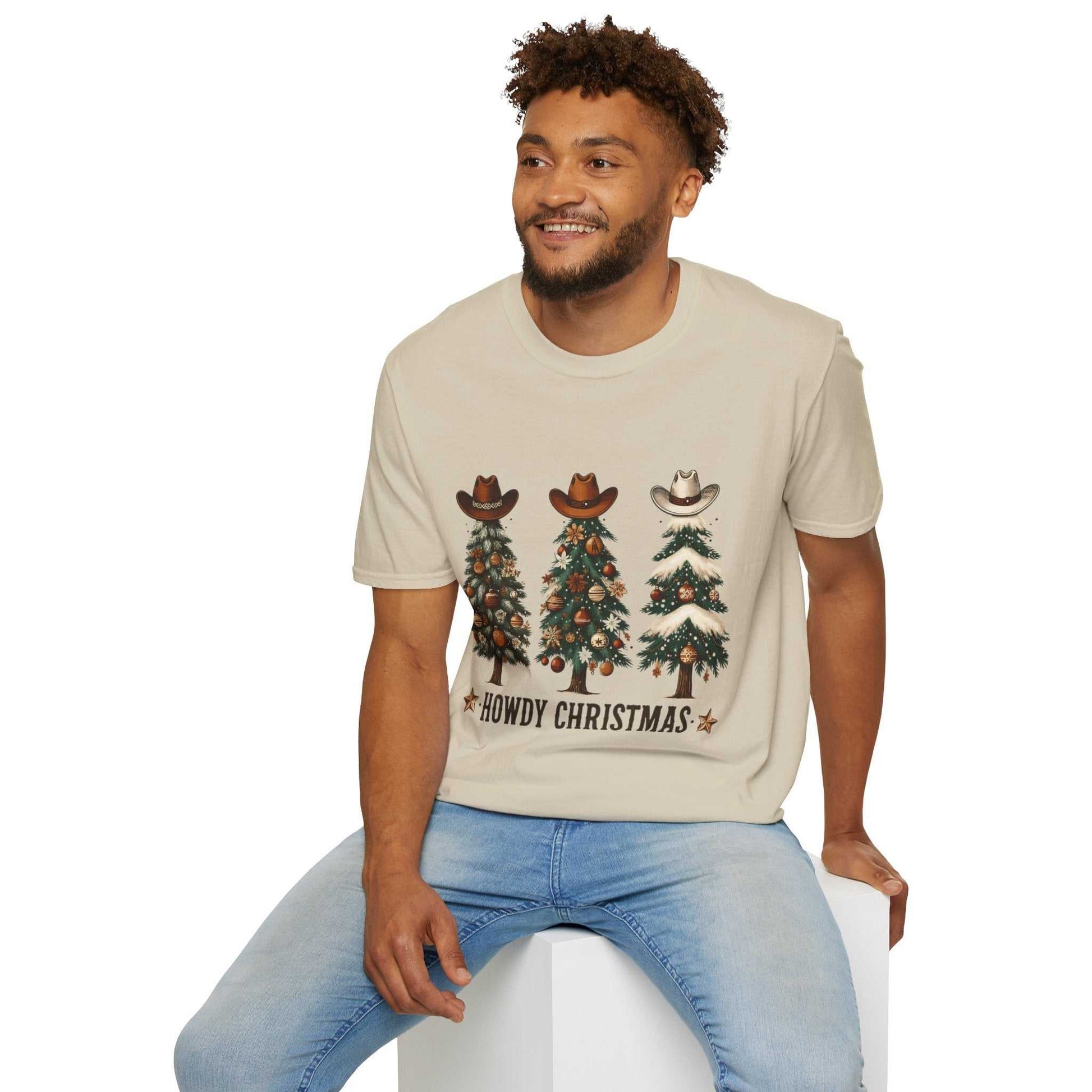 Man wearing beige Howdy Christmas Cowboy Tree T-shirt with decorated Christmas trees topped with cowboy hats and text "Howdy Christmas"