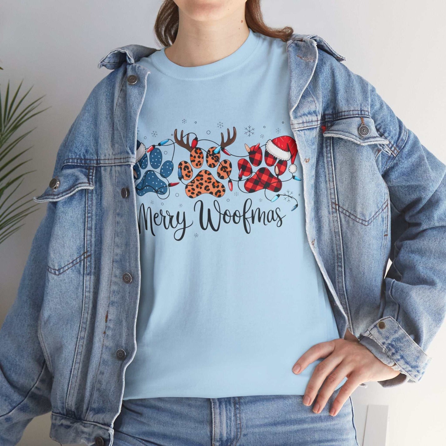 Woman wearing light blue Merry Woofmas Christmas tee with festive paw print design featuring buffalo plaid, leopard spots, Christmas lights, Santa hats, and antlers under denim jacket