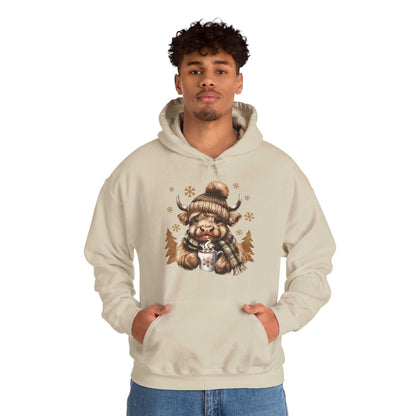 Man wearing beige Highland Cow Hoodie with cozy winter design of Highland cow in knit hat and scarf holding hot chocolate mug