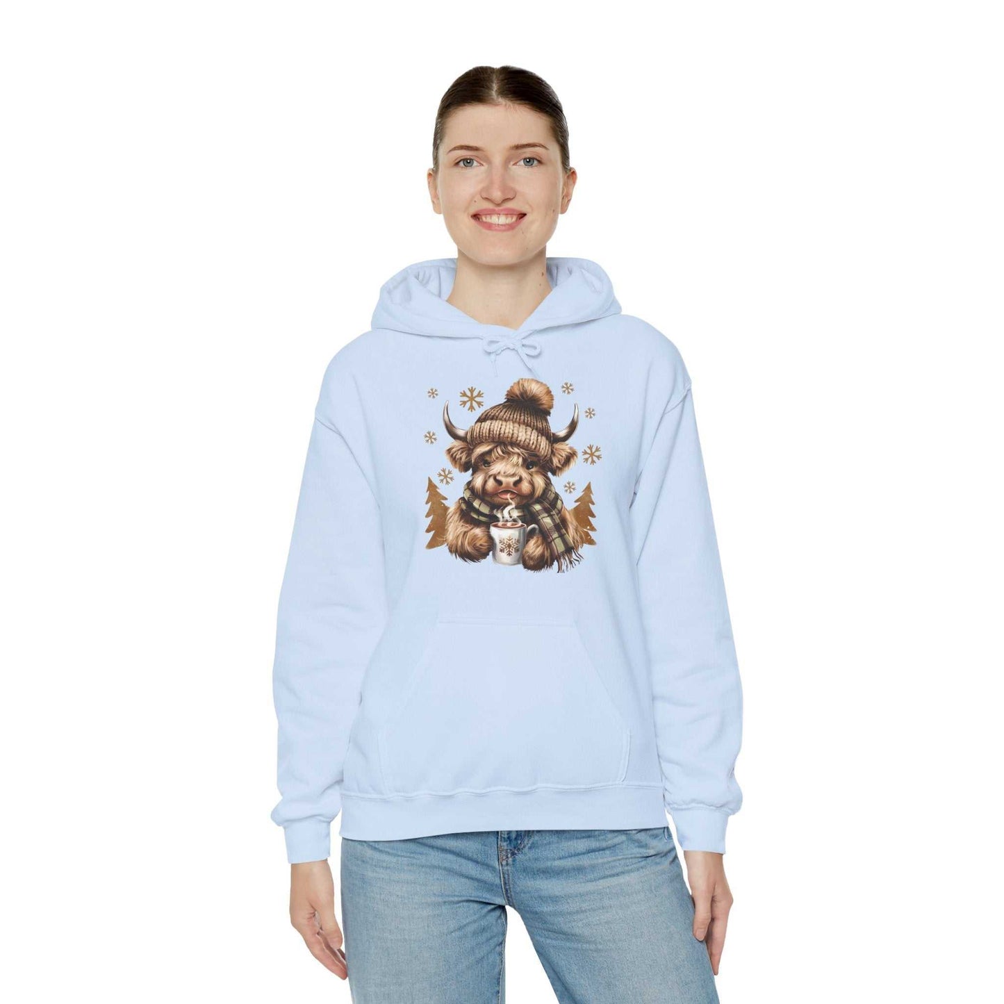 Woman wearing light blue Highland Cow Hoodie with cozy winter design of Highland Cow in hat and scarf holding hot chocolate