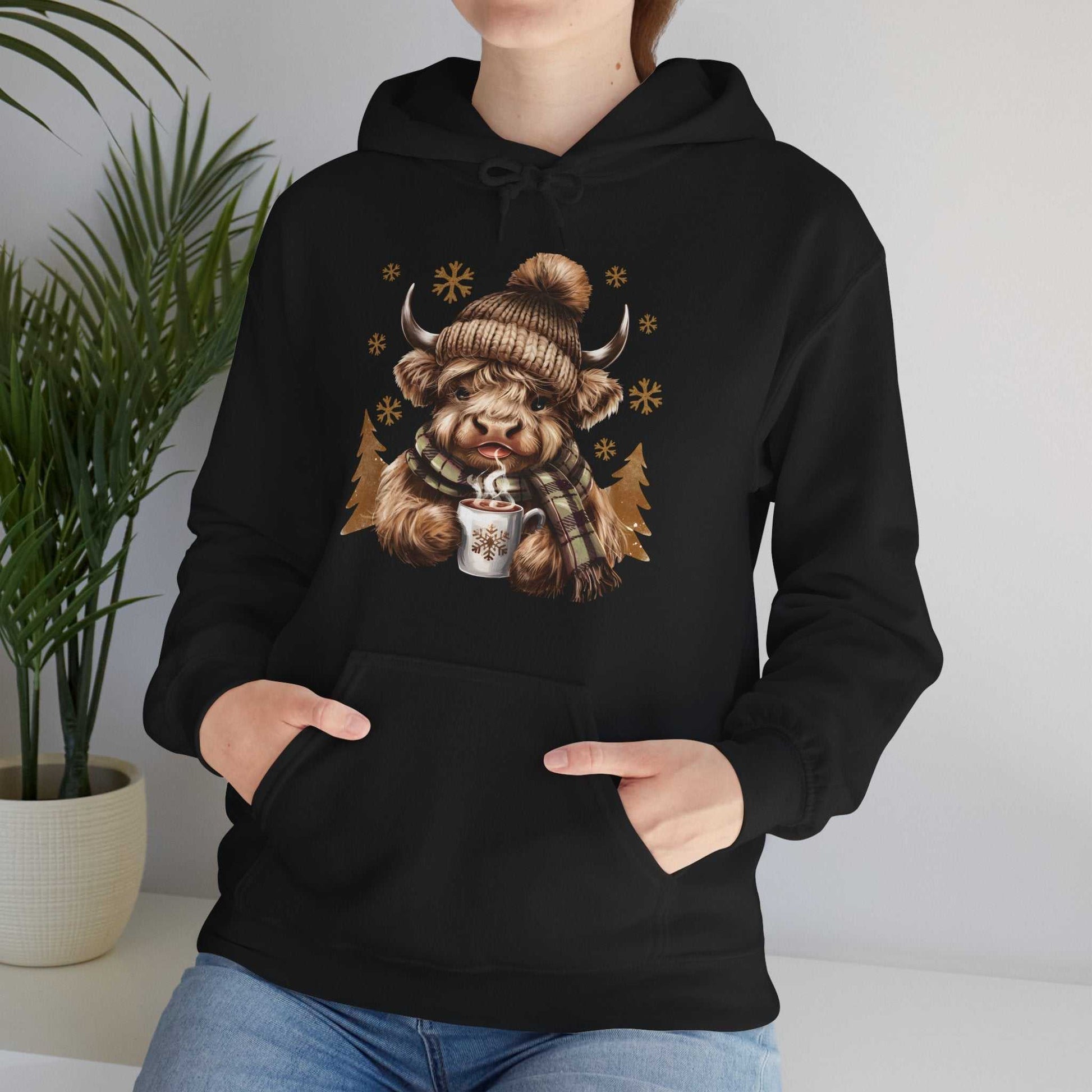 Black Highland Cow hoodie with cozy winter design of cow in knit hat and scarf holding hot chocolate mug