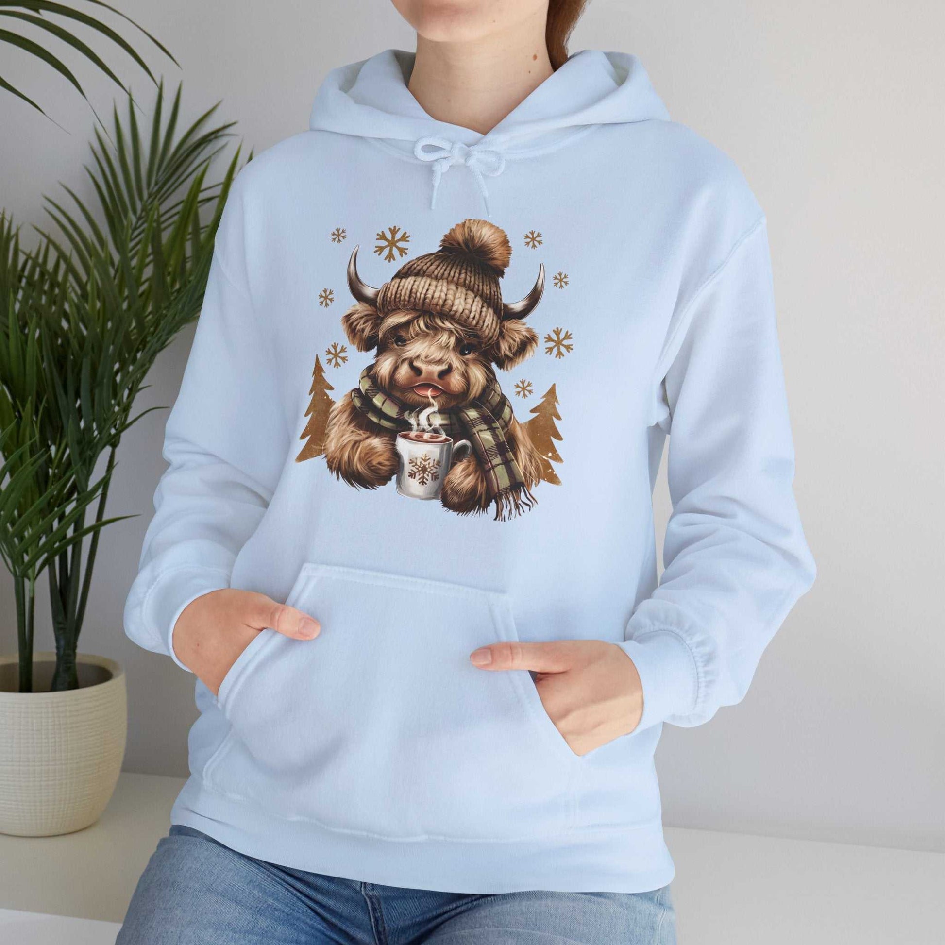 Light blue Highland Cow hoodie with cozy winter cow wearing a wool hat and scarf holding hot chocolate mug with snowflake design.