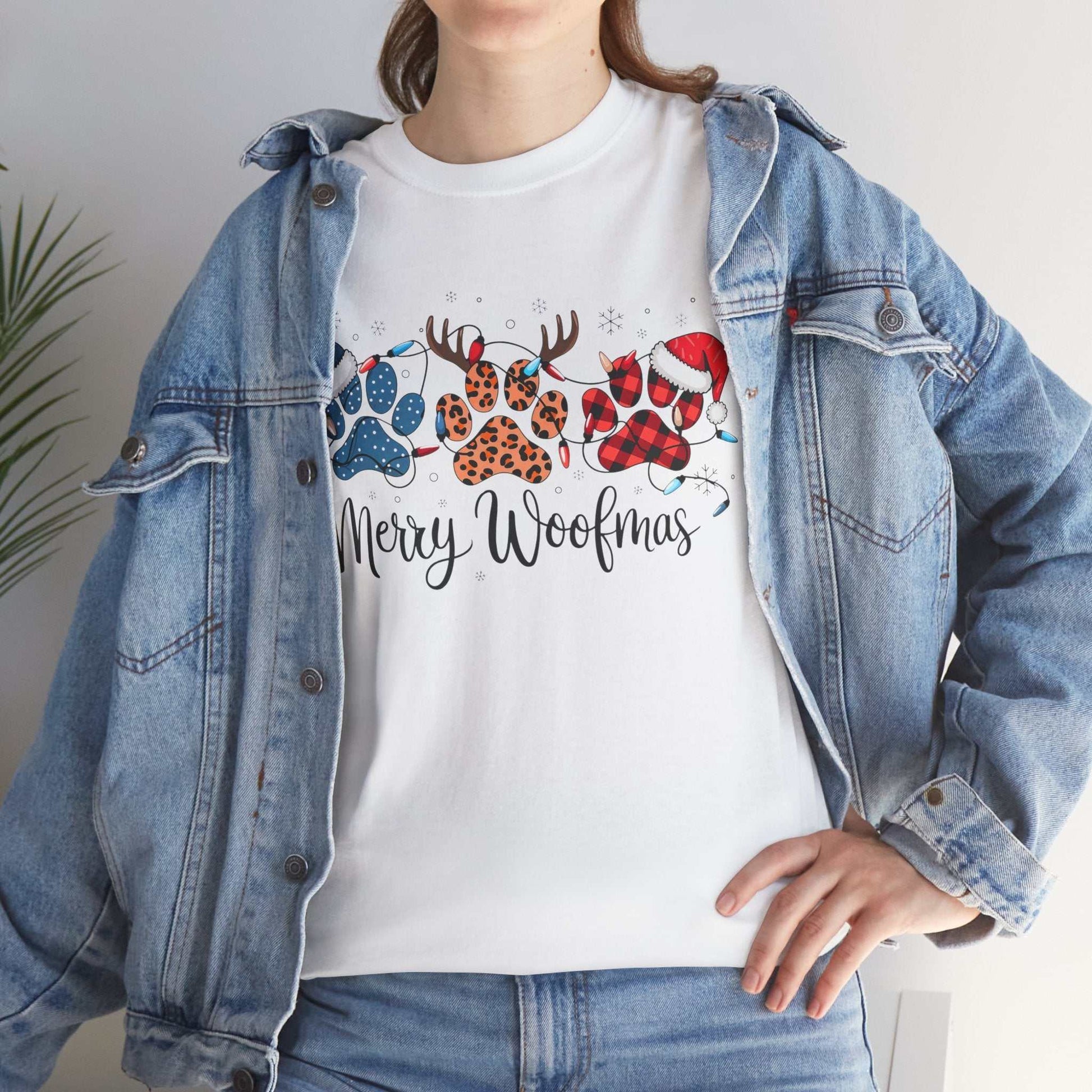 Woman wearing white Merry Woofmas Christmas tee with paw prints in buffalo plaid, leopard spots, Christmas lights, and Santa hats under denim jacket
