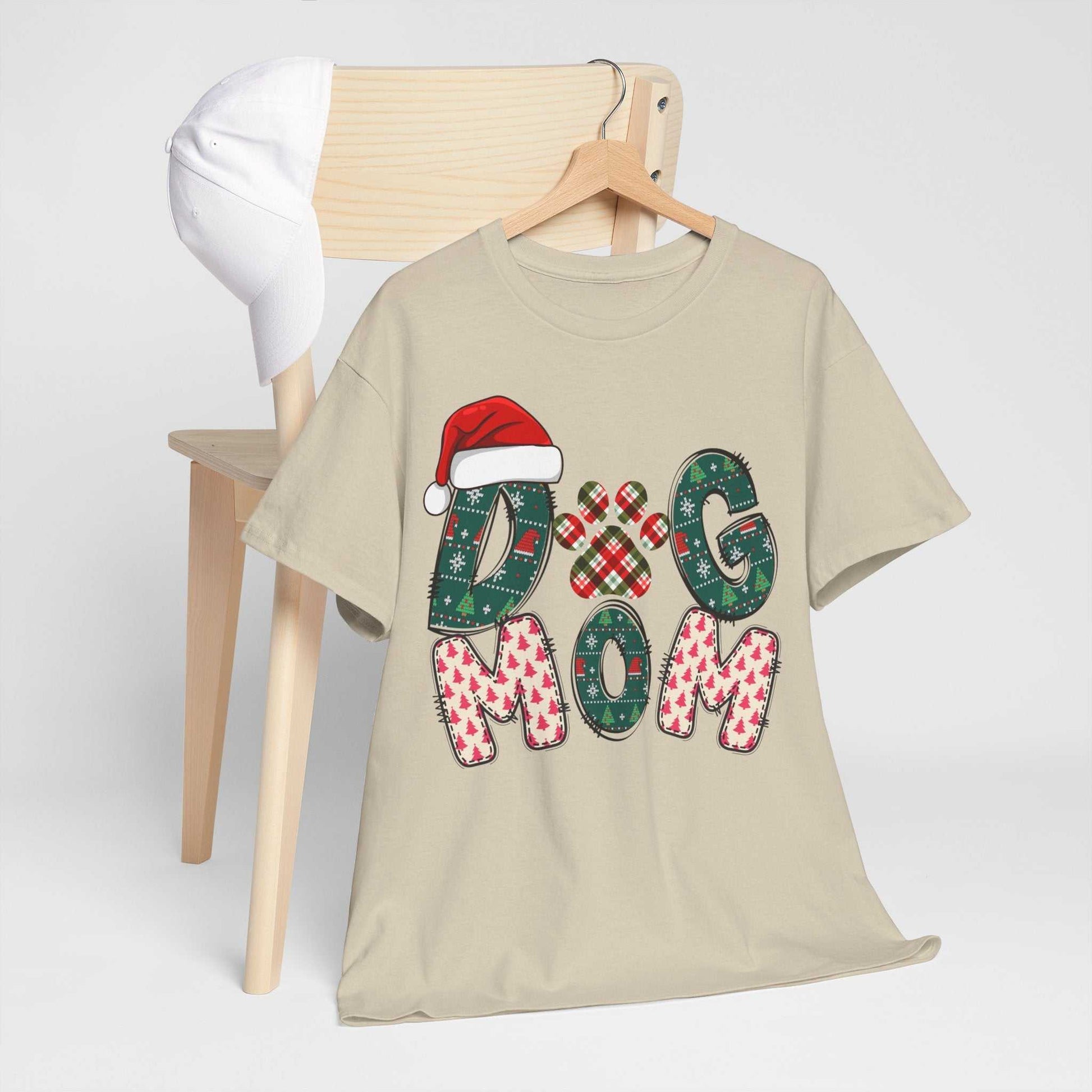 Beige Christmas Dog Mom tee with festive lettering and Santa hat, displayed on hanger by wooden chair with white cap.