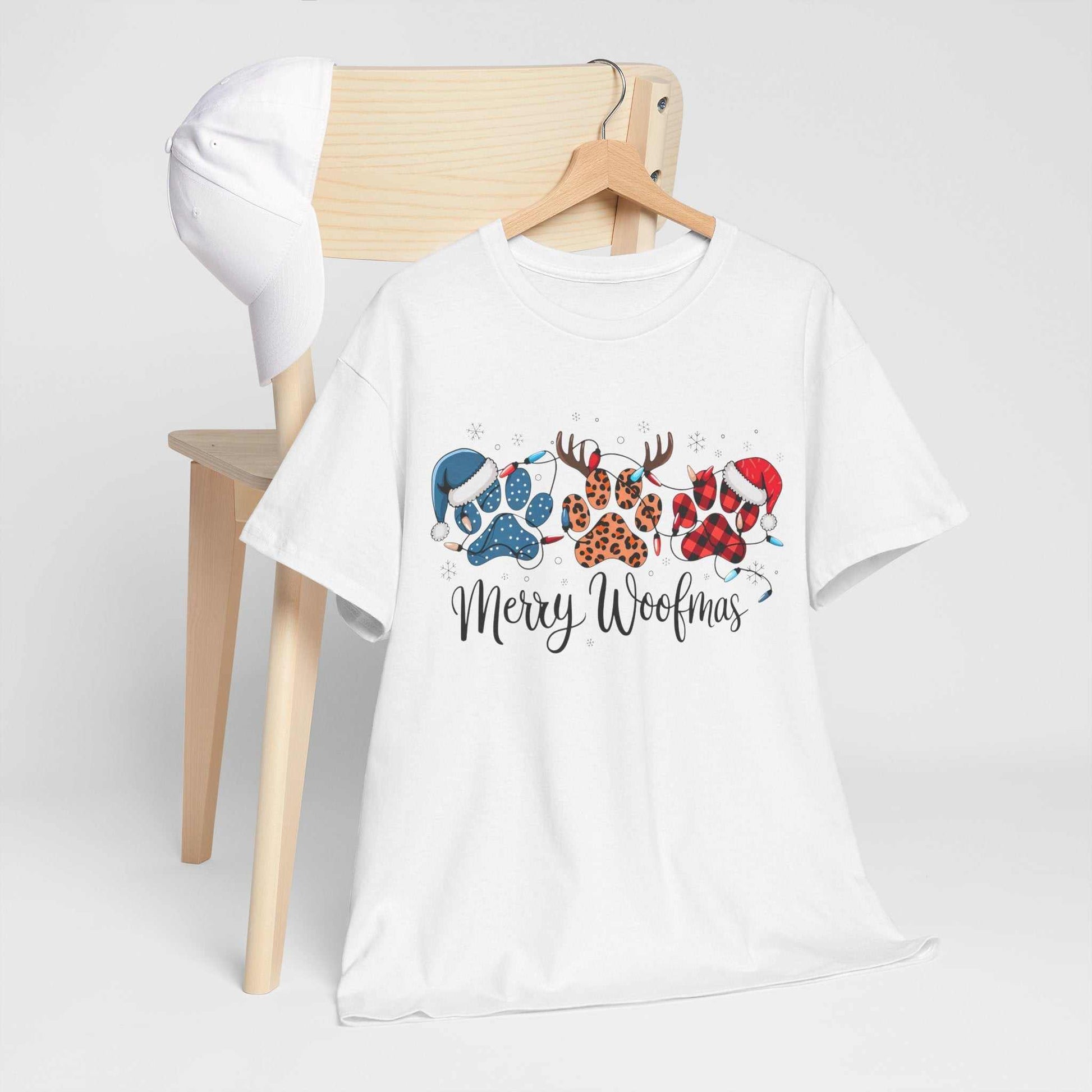 White Merry Woofmas Christmas tee with festive paw prints in buffalo plaid, leopard spots, and Santa hats, displayed on a hanger with a white cap on wooden chair