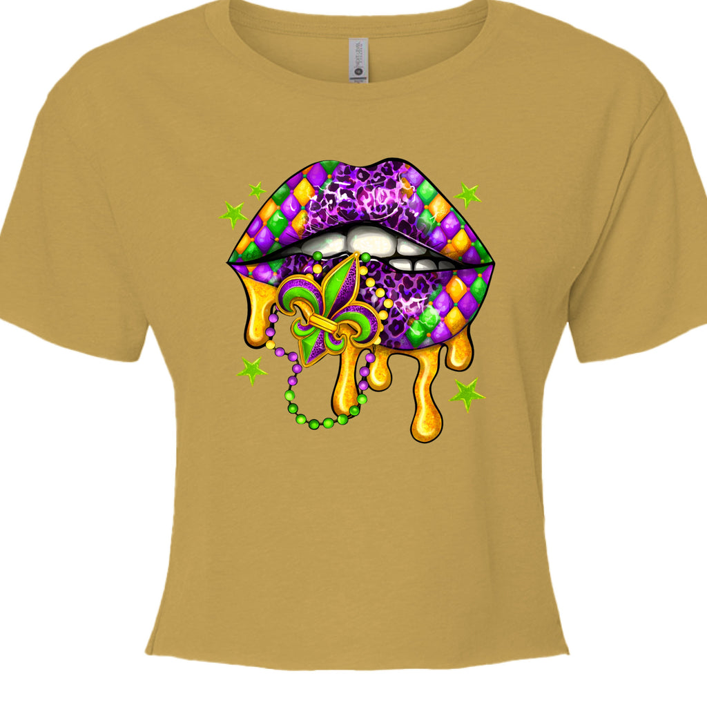 An antique gold, cropped Mardi Gras Lips Baby Tee featuring a large, vibrant graphic of a pair of lips decorated in Mardi Gras themes. The top lip features a purple, gold, and green diamond harlequin pattern, while the bottom lip has a purple leopard print. Gold liquid "drips" from the mouth, which holds a decorative Fleur-de-lis and a string of Mardi Gras beads. 
