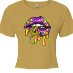 An antique gold, cropped Mardi Gras Lips Baby Tee featuring a large, vibrant graphic of a pair of lips decorated in Mardi Gras themes. The top lip features a purple, gold, and green diamond harlequin pattern, while the bottom lip has a purple leopard print. Gold liquid "drips" from the mouth, which holds a decorative Fleur-de-lis and a string of Mardi Gras beads. 