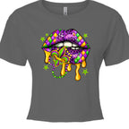 A charcoal grey, cropped Mardi Gras Lips Baby Tee featuring a large, vibrant graphic of a pair of lips decorated in Mardi Gras themes. The top lip features a purple, gold, and green diamond harlequin pattern, while the bottom lip has a purple leopard print. Gold liquid "drips" from the mouth, which holds a decorative Fleur-de-lis and a string of Mardi Gras beads. 