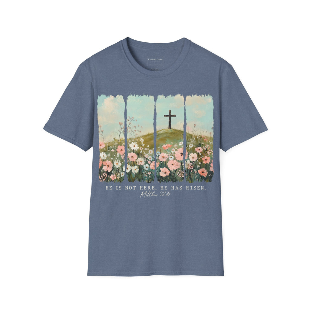 Heather Indigo Easter He Is Risen - Floral Cross Christian Tee featuring a painterly, four-panel graphic of a serene spring landscape. A wooden cross stands on a green hilltop overlooking a field of pink, white, and red wildflowers under a soft blue sky. Below the artwork, the text reads 'HE IS NOT HERE. HE HAS RISEN.' followed by the scripture 'Matthew 28:6' in elegant cursive.
