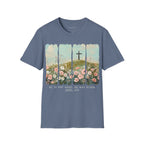 Heather Indigo Easter He Is Risen - Floral Cross Christian Tee featuring a painterly, four-panel graphic of a serene spring landscape. A wooden cross stands on a green hilltop overlooking a field of pink, white, and red wildflowers under a soft blue sky. Below the artwork, the text reads 'HE IS NOT HERE. HE HAS RISEN.' followed by the scripture 'Matthew 28:6' in elegant cursive.