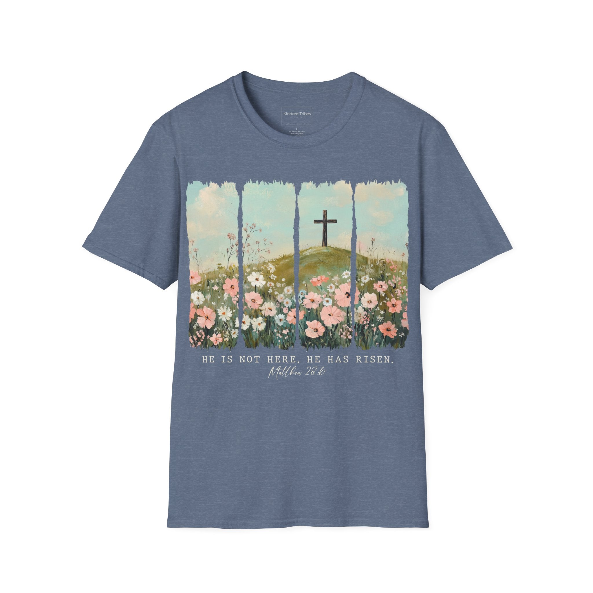 Heather Indigo Easter He Is Risen - Floral Cross Christian Tee featuring a painterly, four-panel graphic of a serene spring landscape. A wooden cross stands on a green hilltop overlooking a field of pink, white, and red wildflowers under a soft blue sky. Below the artwork, the text reads 'HE IS NOT HERE. HE HAS RISEN.' followed by the scripture 'Matthew 28:6' in elegant cursive.