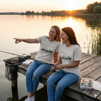 Two women sitting on a dock fishing wearing an ice grey Best Friends Fishin' In The Dark tshirt with a black and white hand sketched image of two best friends in a fishing boat fishing in the moonlight with the saying 'You and Me Goin' Fishin' In The Dark'.