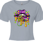 A stonewashed denim color, cropped Mardi Gras Lips Baby Tee featuring a large, vibrant graphic of a pair of lips decorated in Mardi Gras themes. The top lip features a purple, gold, and green diamond harlequin pattern, while the bottom lip has a purple leopard print. Gold liquid "drips" from the mouth, which holds a decorative Fleur-de-lis and a string of Mardi Gras beads. 