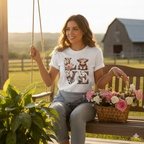 Woman sitting on a porch swing with a basket of roses, in a scenic farm setting, wearing a White t-shirt with a baby highland cow  and floral highlights around wooden and cow hide letters spelling 'LOVE'.