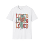 White Loved Cross Heart T-Shirt featuring a large, colorful graphic on the front. The design consists of the word "LOVED" repeated three times in a stacked, chunky block font. The letters are filled with various patterns, including solid colors, stripes, and small hearts in shades of red, sage green, and tan. In the center of the text is a distressed, teal-colored cross with a large, hand-drawn red heart layered over the middle. Below the main graphic, the scripture reference "John 3:16" is written.