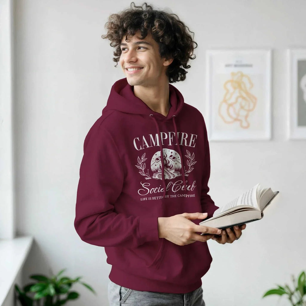 Young man wearing maroon Campfire Social Club cozy hoodie with graphic design, holding an open book indoors