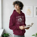 Young man wearing maroon Campfire Social Club cozy hoodie with graphic design, holding an open book indoors